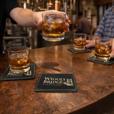 Custom leather whiskey coaster on a distillery bar with a glass being placed above it, showcasing personalized barware and branded bar accessories