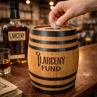 Engraved Larceny Fund oak barrel piggy bank with coin slot on a distillery bar as a hand drops a coin in, showcasing branded distillery gift shop merchandise